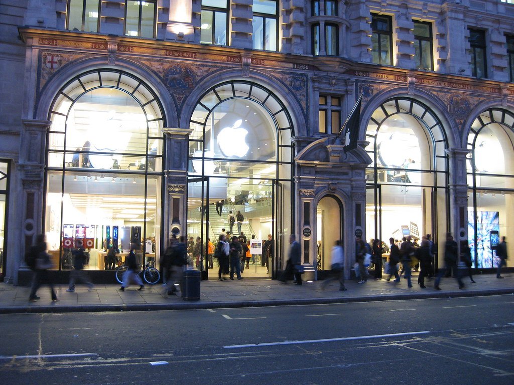 An illuminated Apple Store storefront with the Apple logo, at dusk